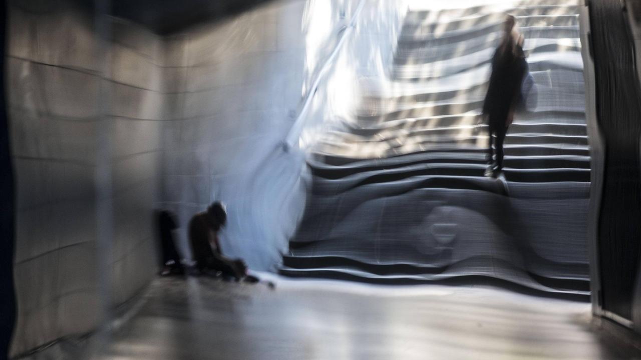 Das Bild zeigt einen Obdachlosen, der in der Fußgängerunterführung sitzt, die unter dem Kaiser-Friedrich-Ring zum Hauptbahnhof Wiesbaden führt. Ein Passant läuft vorbei. Die Szene spiegelt sich verzerrt in der spiegelnden Wandverkleidung der Unterführung. Das Bild zeigt einen Obdachlosen, der in der Fußgängerunterführung sitzt, die unter dem Kaiser-Friedrich-Ring zum Hauptbahnhof Wiesbaden führt. Ein Passant läuft vorbei. Die Szene spiegelt sich verzerrt in der spiegelnden Wandverkleidung der Unterführung.