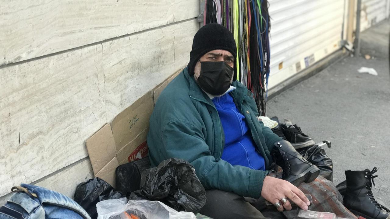 Ein älterer Mann sitzt mit Maske, Mütze und Schuhputzutensilien auf dem Boden, mit vielen Tüten und Taschen um sich herum. Ein älterer Mann sitzt mit Maske, Mütze und Schuhputzutensilien auf dem Boden, mit vielen Tüten und Taschen um sich herum.