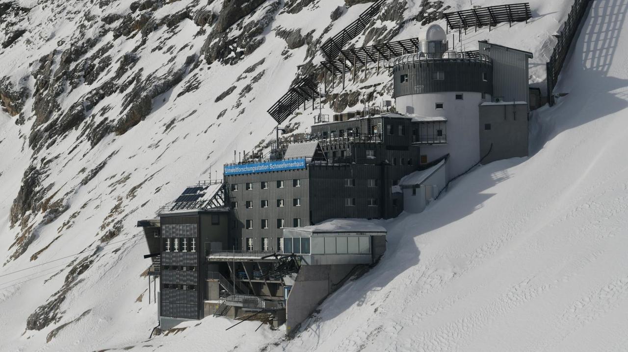 Ein Farbfoto zeigt einen terrassierten, grau verkleideten Gebäudekomplex inmitten eines steil abfallenden, schneebedeckten Felsmassivs Ein Farbfoto zeigt einen terrassierten, grau verkleideten Gebäudekomplex inmitten eines steil abfallenden, schneebedeckten Felsmassivs