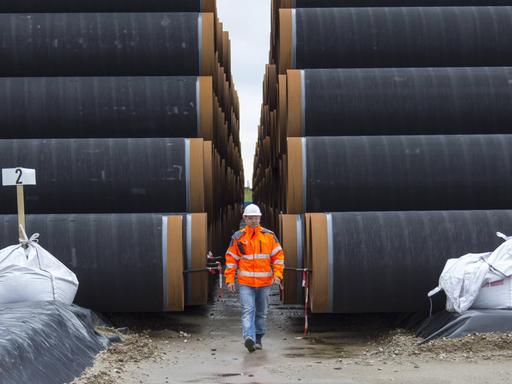Ein Mann mit Schutzweste und Helm läuft im Fährhafen Sassnitz-Mukran auf Rügen zwischen zwei zu Türmen gestapelten Rohren für die Gaspipeline Nord Stream 2 hindurch.