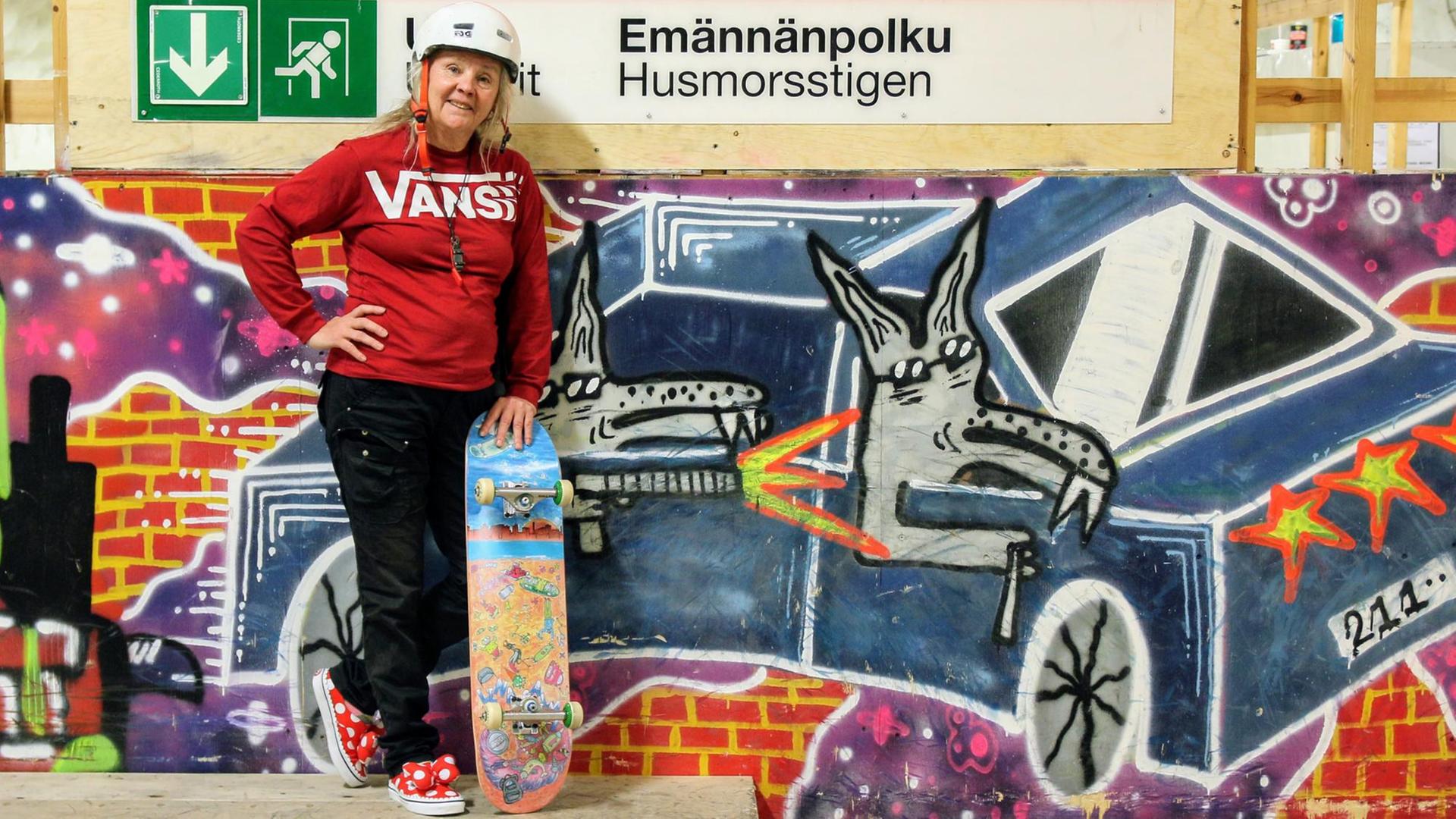 Lena Salmi steht mit rotem Pulli und typischem Skateboard-Helm vor einer bunt besprühten Graffiti-Wand, während ein farbenfrohes Skateboard lässig neben ihr lehnt.