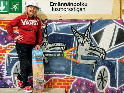 Lena Salmi steht mit rotem Pulli und typischem Skateboard-Helm vor einer bunt besprühten Graffiti-Wand, während ein farbenfrohes Skateboard lässig neben ihr lehnt.