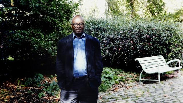 Achille Mbembe steht in einem blauem Anzug in einem Pariser Park vor grünem Gebüsch und einer weißen Bank.