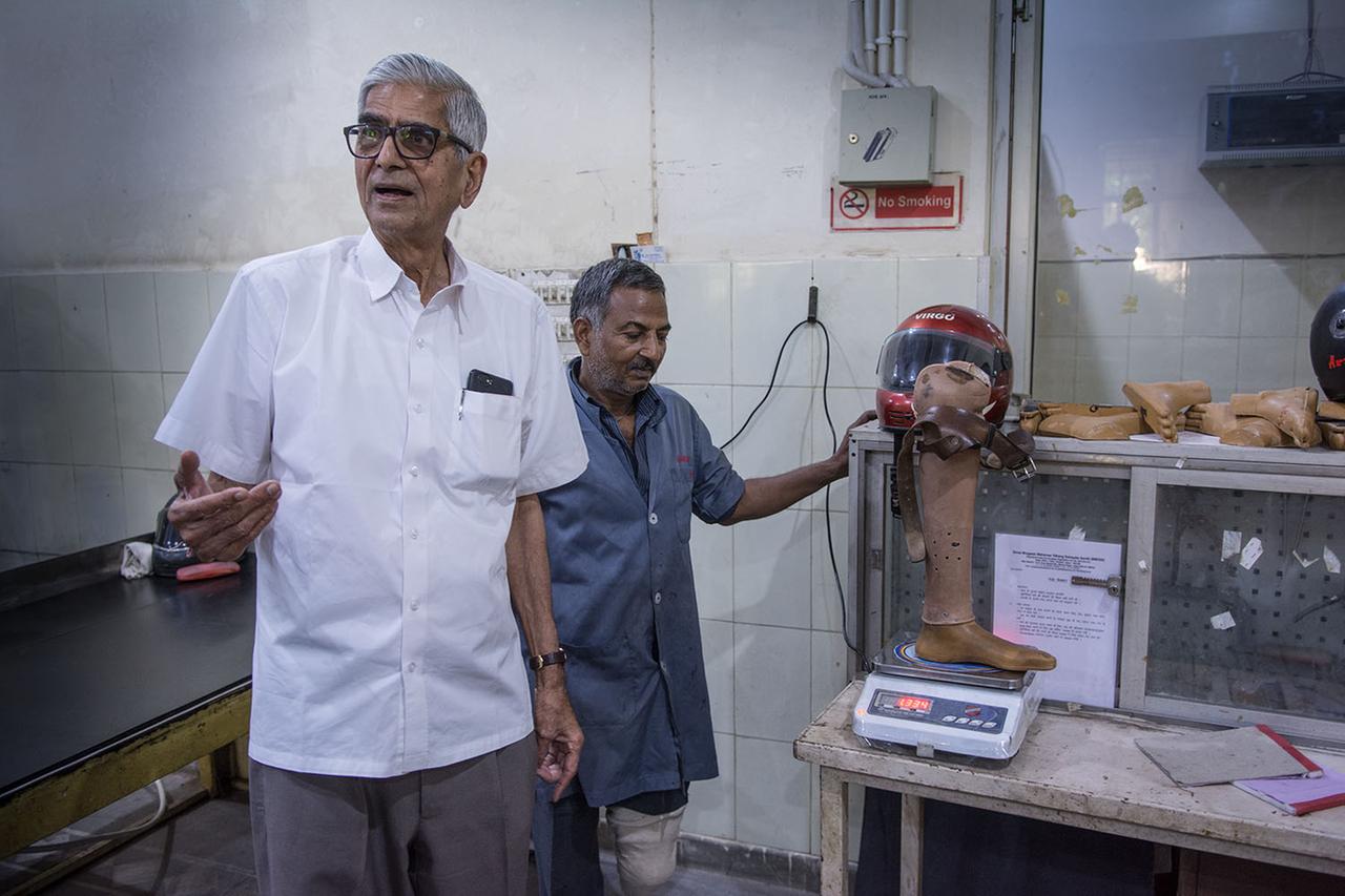 Dr. Mehta steht mit einem Mitarbeiter in der Werkstatt, beide schauen in die Kamera, im Hintergrund eine Prothese auf einer Waage Dr. Mehta steht mit einem Mitarbeiter in der Werkstatt, beide schauen in die Kamera, im Hintergrund eine Prothese auf einer Waage