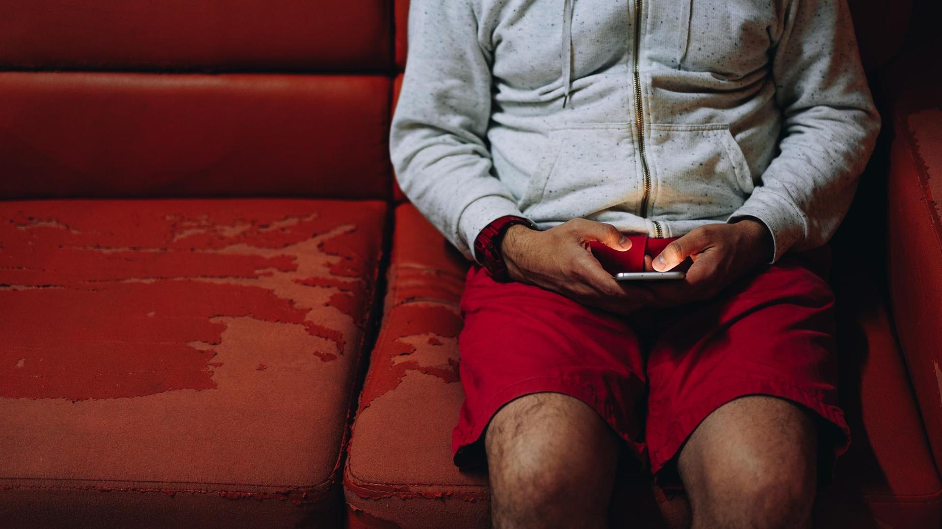 Ein Mann sitzt auf einer alten roten Couch und schaut auf sein Handy. Ein Mann sitzt auf einer alten roten Couch und schaut auf sein Handy.