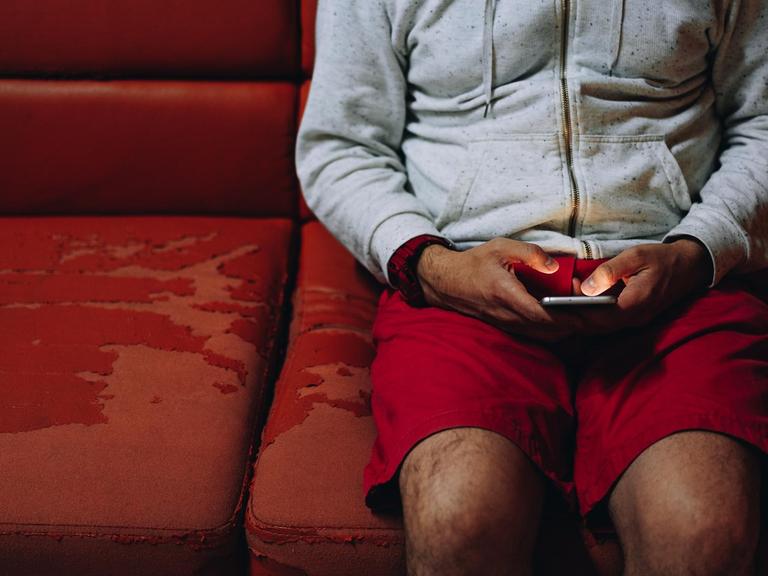 Ein Mann sitzt auf einer alten roten Couch und schaut auf sein Handy.