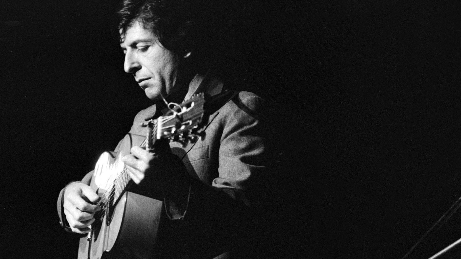 Schwarzweißfoto von Leonard Cohen, der nachdenklich und Gitarre spielend auf einer Bühne steht Schwarzweißfoto von Leonard Cohen, der nachdenklich und Gitarre spielend auf einer Bühne steht