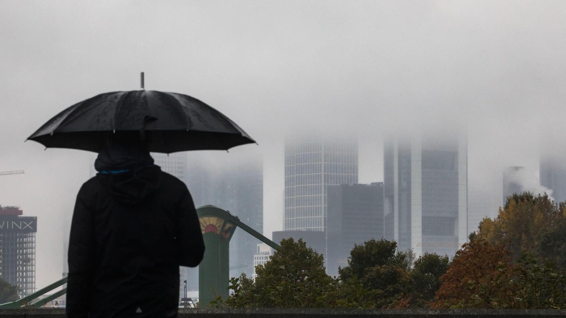 Ein Mann geht am 24.10.2016 in Frankfurt am Main (Hessen) mit aufgespanntem Schirm an der regnerischen Kulisse der Stadt vorbei. Die Spitzen der Hochhäuser verschwinden im Nebel. Ein Mann geht am 24.10.2016 in Frankfurt am Main (Hessen) mit aufgespanntem Schirm an der regnerischen Kulisse der Stadt vorbei. Die Spitzen der Hochhäuser verschwinden im Nebel.