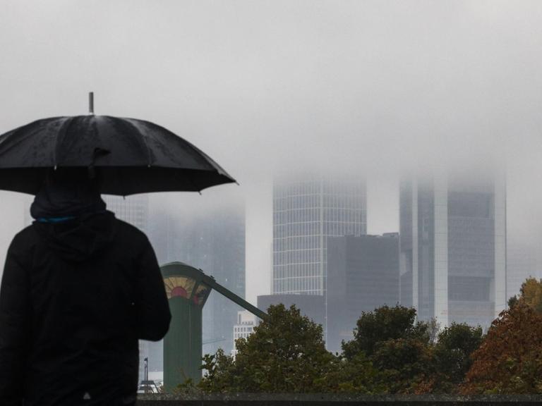 Ein Mann geht am 24.10.2016 in Frankfurt am Main (Hessen) mit aufgespanntem Schirm an der regnerischen Kulisse der Stadt vorbei. Die Spitzen der Hochhäuser verschwinden im Nebel.