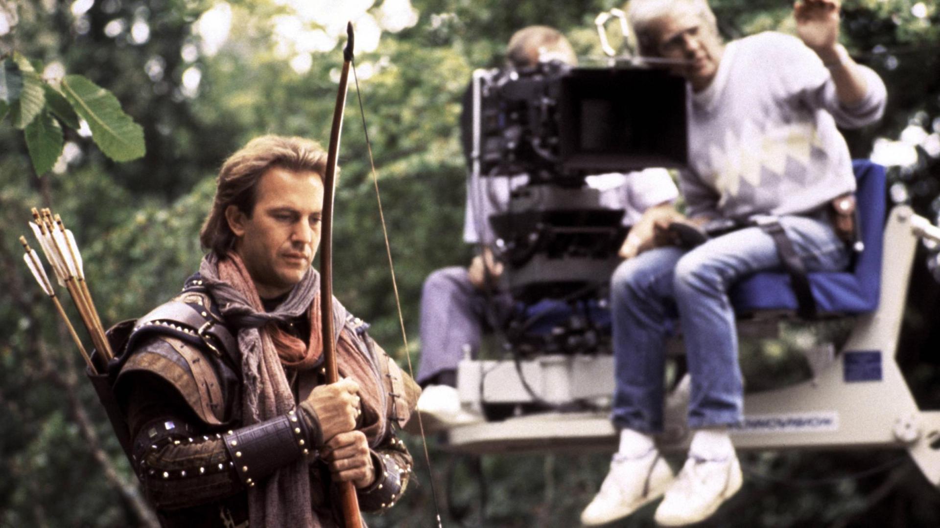 Kevin Costner steht im Robin-Hood-Kostüm samt Bogen vor einem Kamerakran