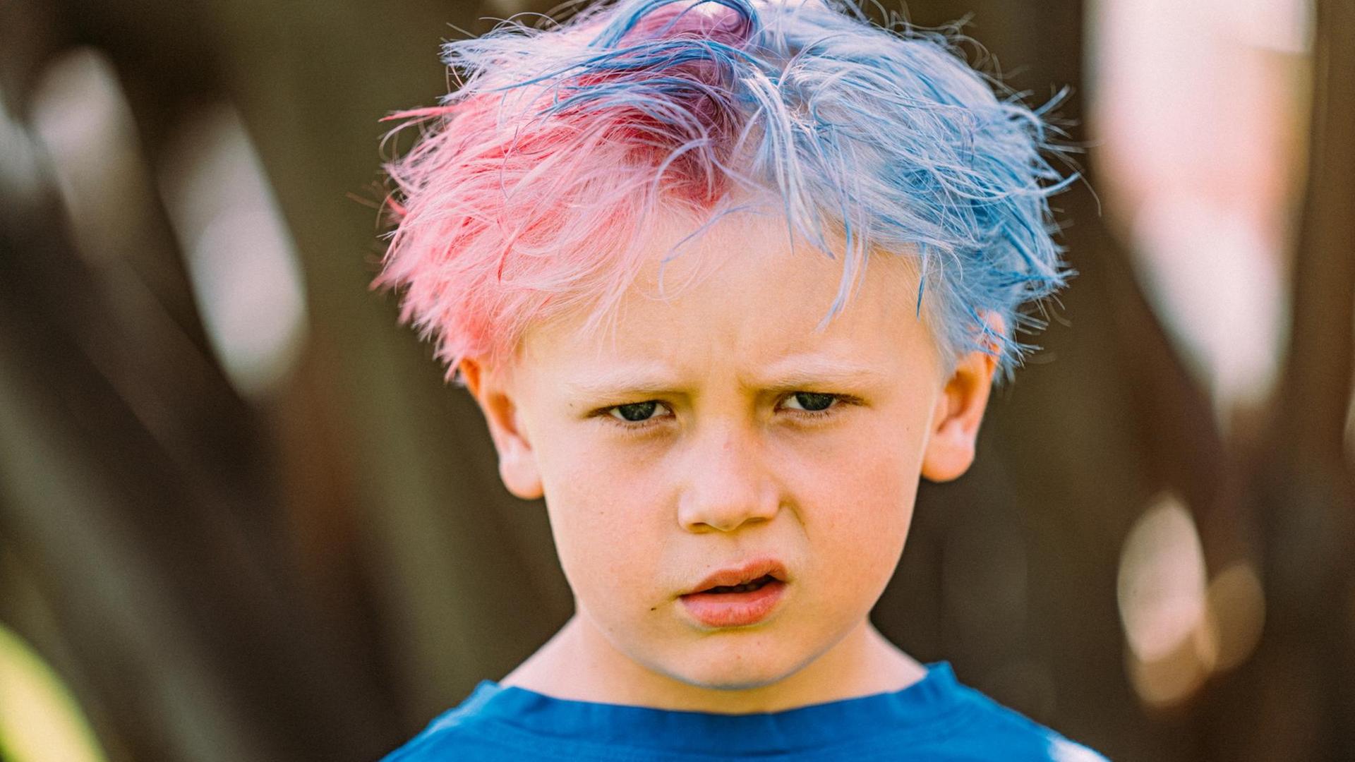 Ein Kind mit zusammengeschobenen Augenbrauen und hochgezogenem Mundwinkel. Die Haare sind zur Hälfte pink und zur Hälfte blau. Ein Kind mit zusammengeschobenen Augenbrauen und hochgezogenem Mundwinkel. Die Haare sind zur Hälfte pink und zur Hälfte blau.