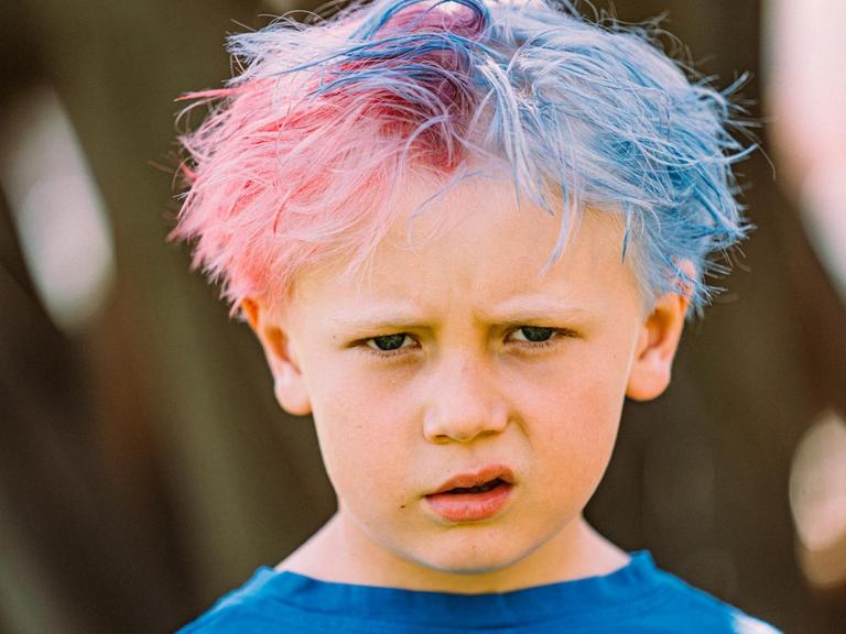 Ein Kind mit zusammengeschobenen Augenbrauen und hochgezogenem Mundwinkel. Die Haare sind zur Hälfte pink und zur Hälfte blau.