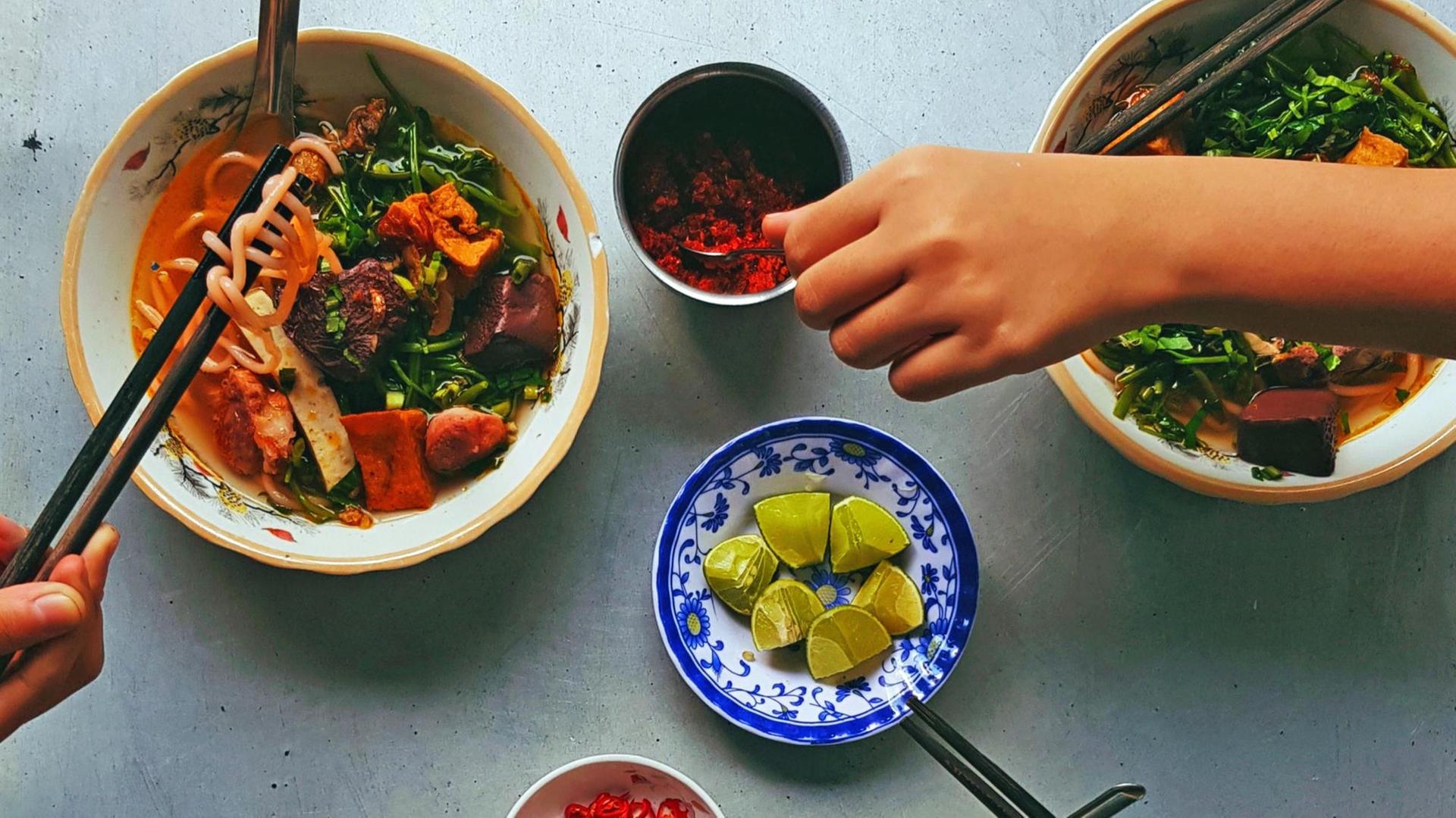 Draufsicht auf verschiedene Schalen mit asiatischem Essen, dazu eine Hand die nach Essen greift.