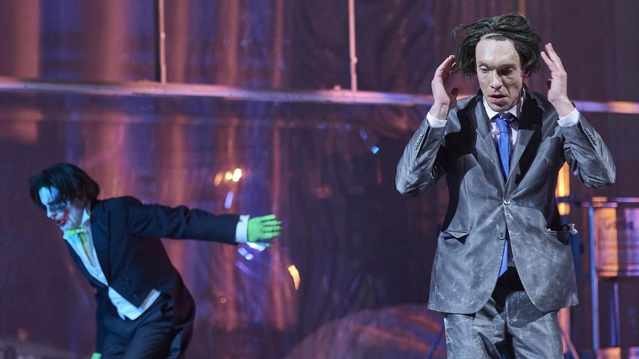 Szenenfoto: Ein Mann im Anzug und mit zerzausten Haaren hebt die Hände Richtung Kopf. Neben ihm steht ein Mann, der aussieht wie die Comic-Figur "Joker". Verwüstung auf der Bühne. Szenenfoto: Ein Mann im Anzug und mit zerzausten Haaren hebt die Hände Richtung Kopf. Neben ihm steht ein Mann, der aussieht wie die Comic-Figur "Joker". Verwüstung auf der Bühne.