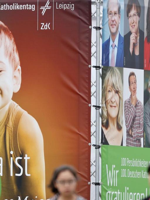 Passanten laufen an einer viereckigen Installation mit Transparenten vorbei. Auf der Vorserseite sieht man das überlebensgroße Bild eines grinsenden Jungen und die Schrift "Seht, da ist der Mensch".