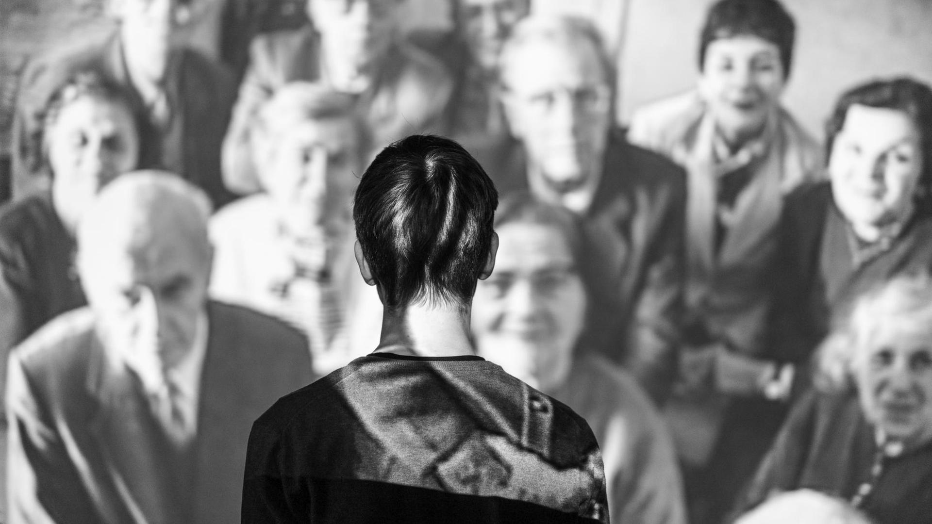 Eine Person steht mit dem Gesicht zur Wand vor einer Videoinstallation auf der viele ältere Menschen zu sehen sind. Eine Person steht mit dem Gesicht zur Wand vor einer Videoinstallation auf der viele ältere Menschen zu sehen sind.