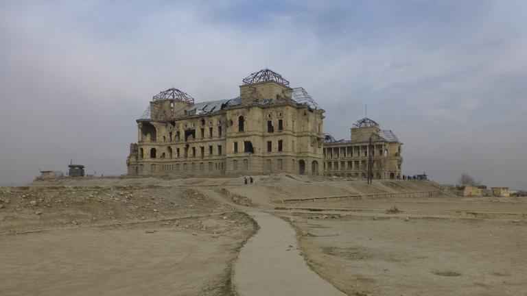 Ruine des Darul-Aman-Palastes: ein rechteckiges pompöses und zerbombtes Gebäude aus der Ferne, eine einsame Straße führt nach dort
