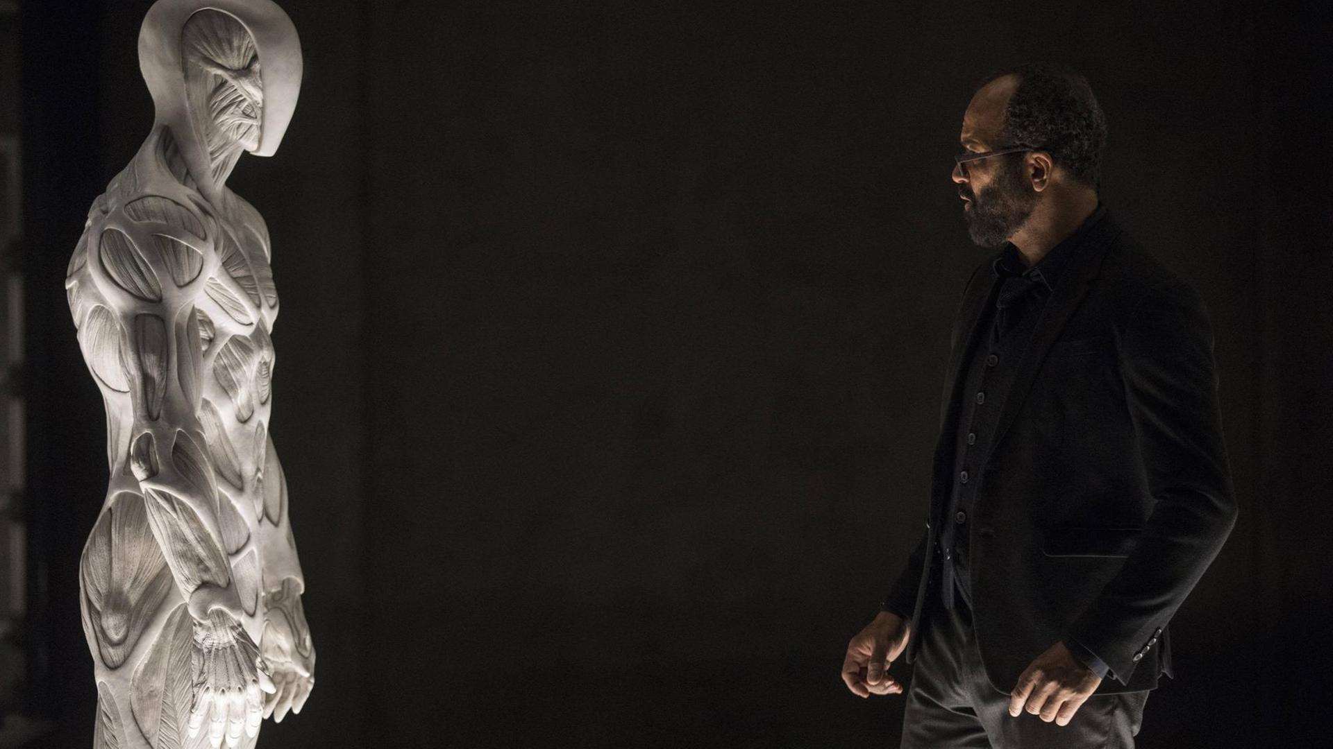 Jeffrey Wright steht vor schwarzem Hintergrund einem alabasterfarbenen künstlichen Menschen gegenüber Jeffrey Wright steht vor schwarzem Hintergrund einem alabasterfarbenen künstlichen Menschen gegenüber