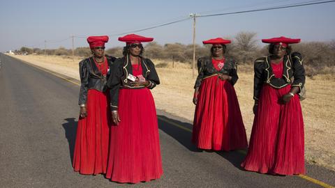 Vier Frauen in ähnlichen roten Kleidern, schwarzer Blouson-Jacke und roter, traditioneller Herero-Kopfbedeckung stehen auf einer leeren Straße.