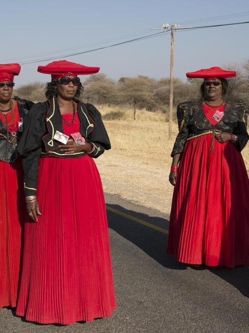 Vier Frauen in ähnlichen roten Kleidern, schwarzer Blouson-Jacke und roter, traditioneller Herero-Kopfbedeckung stehen auf einer leeren Straße.