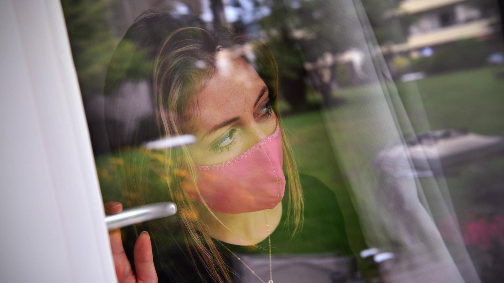 Eine junge Frau mit Schutzmaske steht am Fenster in einem Zimmer und blickt hinaus. Eine junge Frau mit Schutzmaske steht am Fenster in einem Zimmer und blickt hinaus.