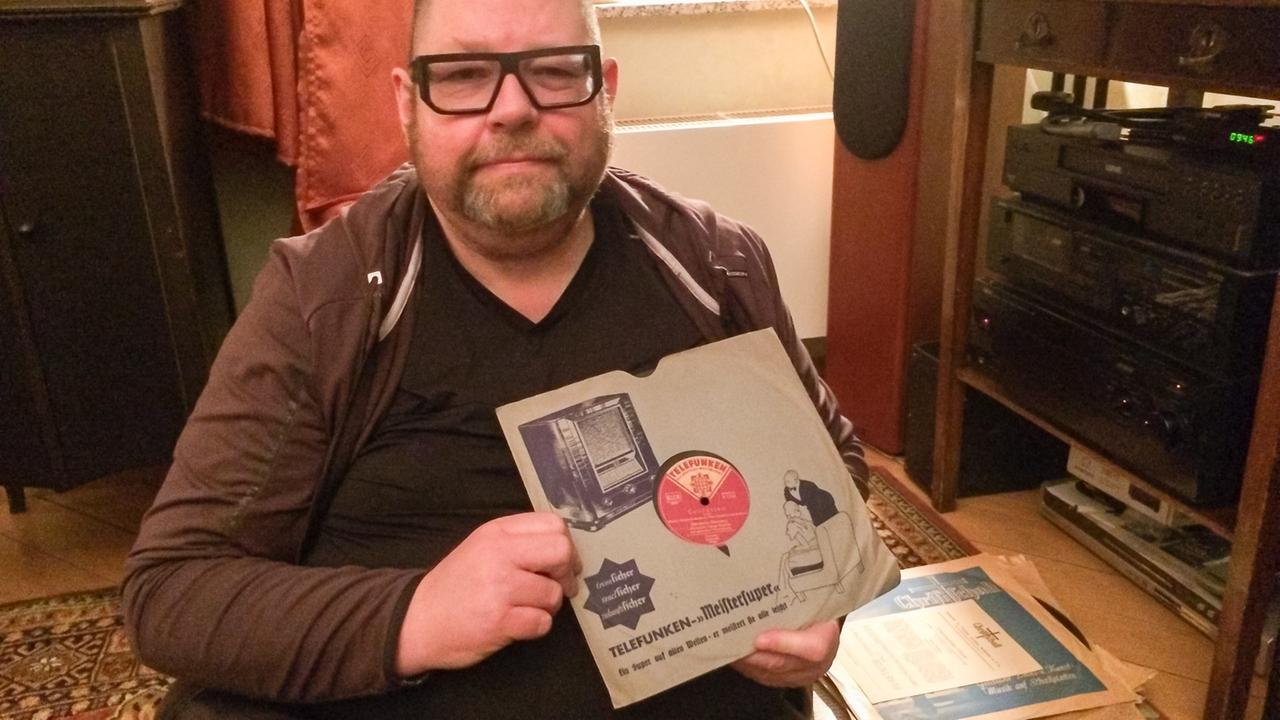 Claus Fischer sitzt auf dem Boden seiner Wohnung und hält eine seiner Schellack-Raritäten mit Radiowerbung in die Kamera. Claus Fischer sitzt auf dem Boden seiner Wohnung und hält eine seiner Schellack-Raritäten mit Radiowerbung in die Kamera.