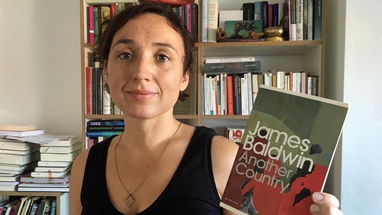Redakteurin Julia Riedhammer steht vor einem Bücherregal und hält das Buch "Another Country" in die Kamera.
