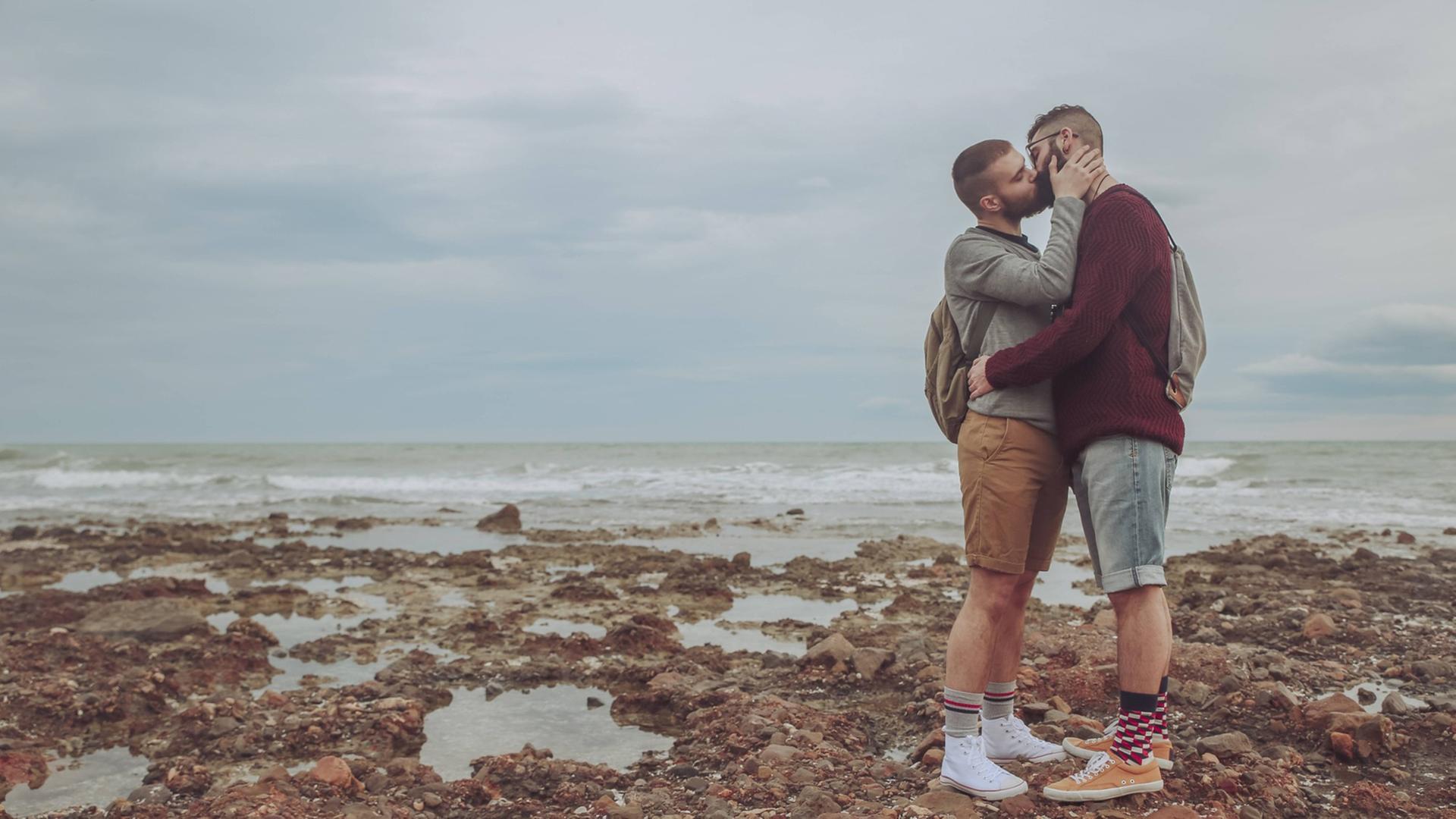Zwei Männer küssen sich auf einem steinigen Strand, im Hintergrund ist das Meer zu sehen. Zwei Männer küssen sich auf einem steinigen Strand, im Hintergrund ist das Meer zu sehen.