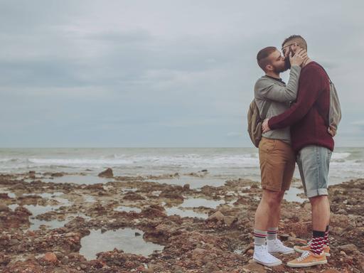 Zwei Männer küssen sich auf einem steinigen Strand, im Hintergrund ist das Meer zu sehen.