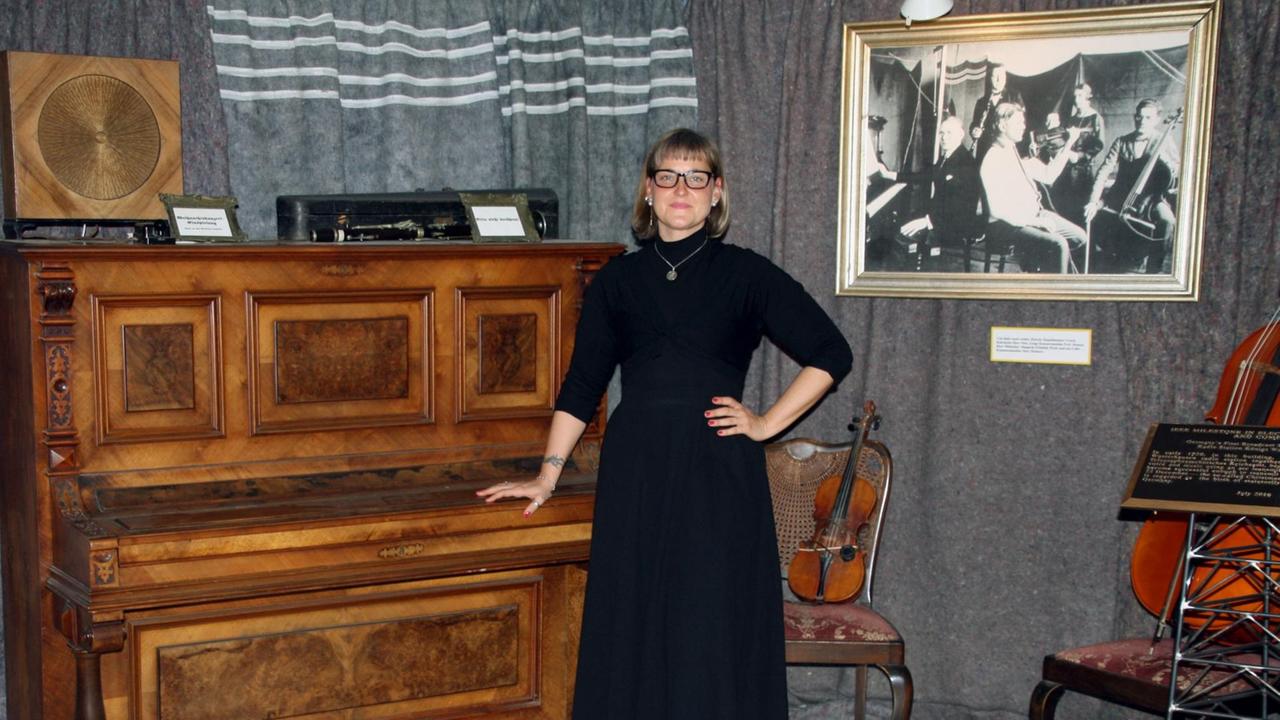 Eine Frau in schwarzem Pullover und langem Rock steht neben einem alten Klavier aus edlem Holz, auf das sie vorsichtig ihre Hand gelegt hat.