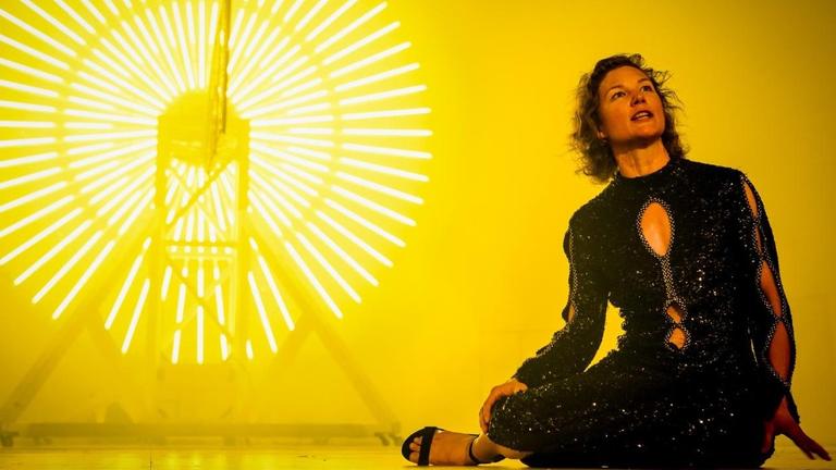 Die Schauspielerin Cordelia Wege in einer Szene des Stücks "Lear" in der Regie von Sebastian Hartmann am Deutschen Theater in Berlin. Sie trägt ein dunkles Kleid und sitzt auf der Bühne. Hinter ihr erstrahlen kreisrunde Leuchtröhren vor einem gelben Hintergrund.