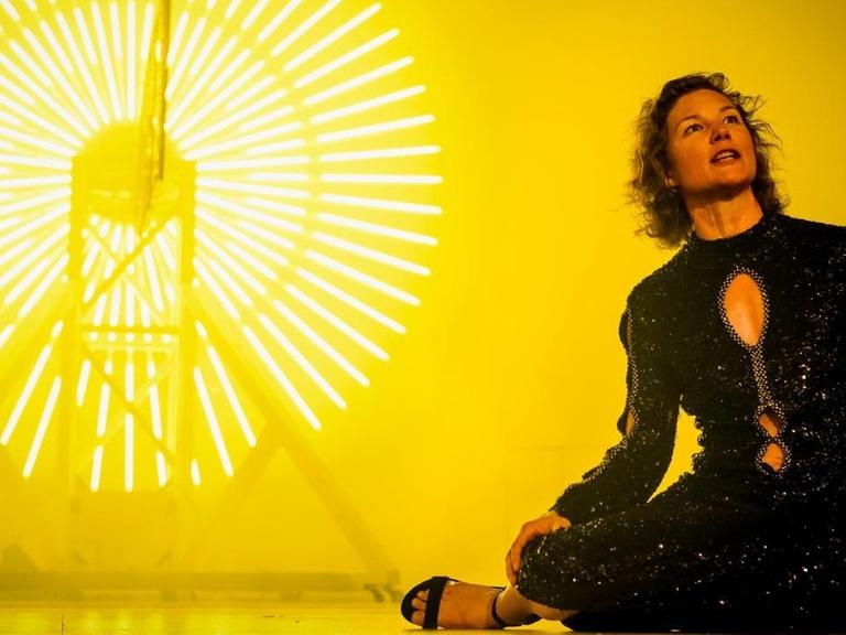 Die Schauspielerin Cordelia Wege in einer Szene des Stücks "Lear" in der Regie von Sebastian Hartmann am Deutschen Theater in Berlin. Sie trägt ein dunkles Kleid und sitzt auf der Bühne. Hinter ihr erstrahlen kreisrunde Leuchtröhren vor einem gelben Hintergrund.