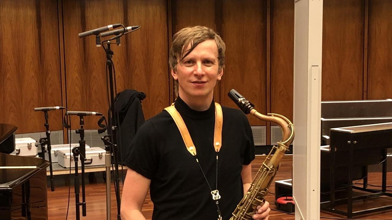 Ein blonder Mann in schwarzem Shirt posiert mit Tenorsaxofon vor holzgetäfelter Wand Ein blonder Mann in schwarzem Shirt posiert mit Tenorsaxofon vor holzgetäfelter Wand