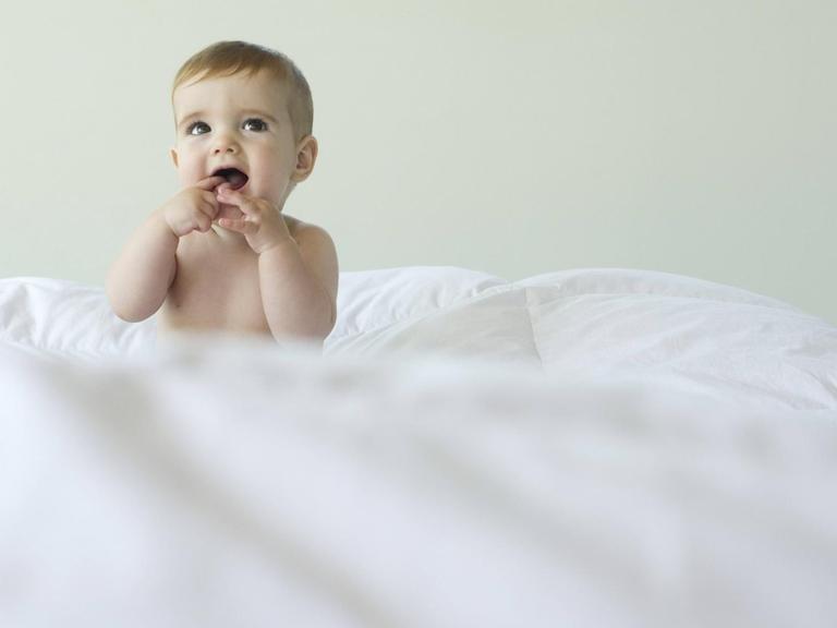 Ein Baby sitzt auf einem Bett und steckt sich die Finger in den Mund.