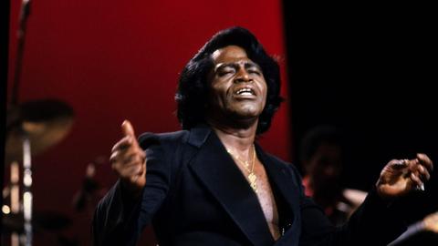 James Brown live auf einem Konzert in den 80er Jahren. Der Musiker trägt ein schwarzes Hemd, der Hintergrund ist in weinrotes Licht getaucht.