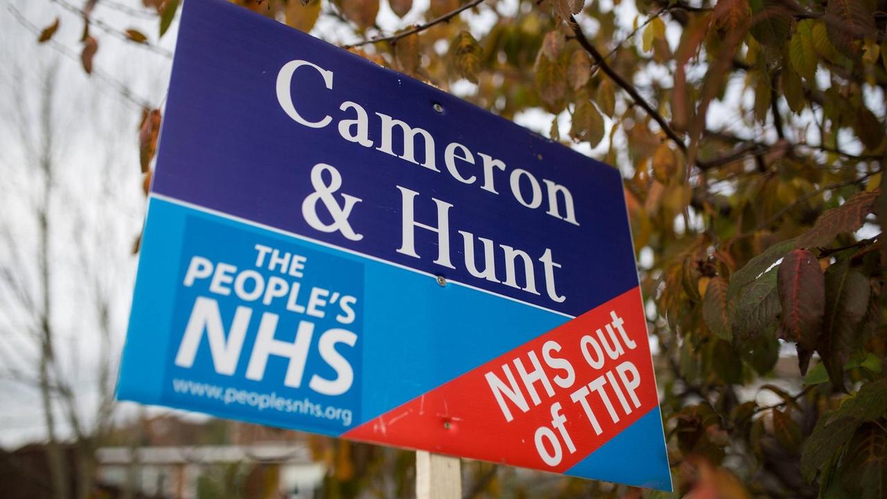 Ein Plakat an einem Stock wird in die Luft gehalten, darauf die Aufschrift: "Cameron & Hunt - The People's NHS - NHS out of TTIP"
