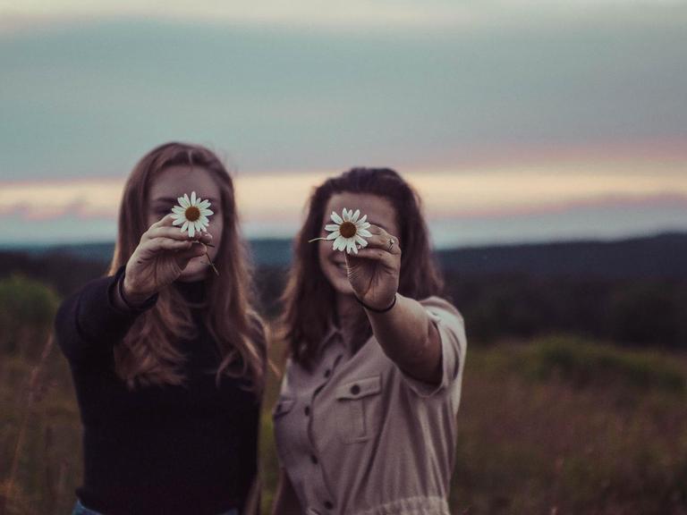 Zwei Frauen stehen nebeneinander und halten sich eine Blume vor das Gesicht.