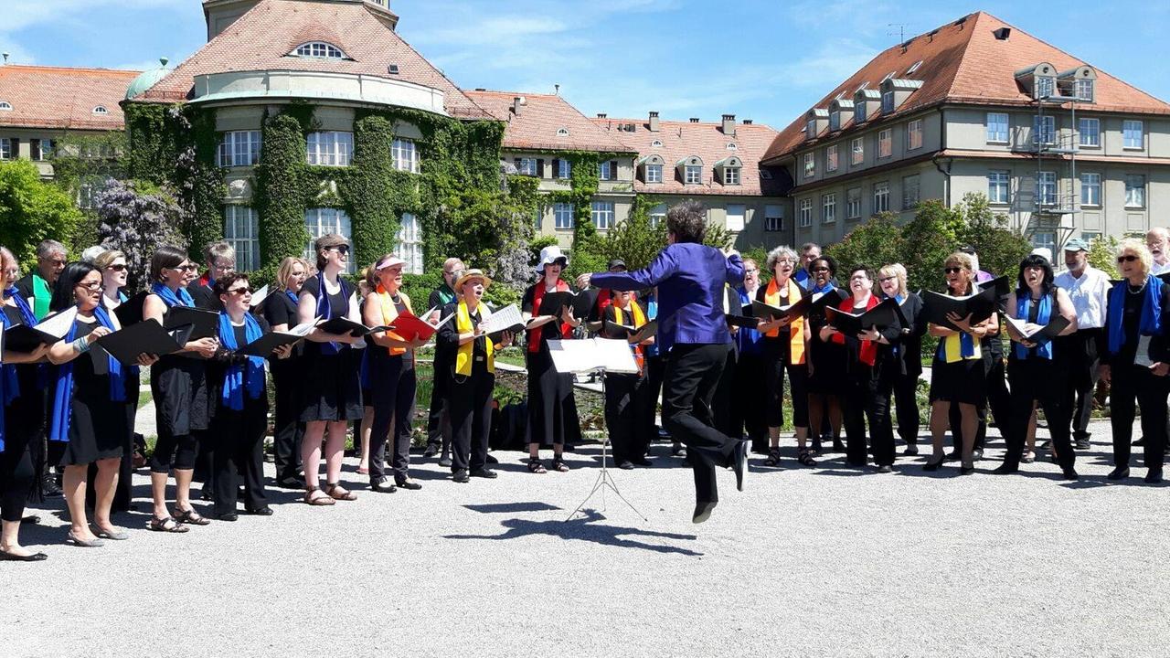 Ein bunt gekleideter Chor ist im Halbkreis zu einem Open Air Konzert in einem Park aufgestellt. Die Leiterin macht einen Hüpfer beim Dirigieren.