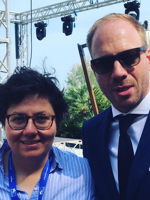 Schauspieler Johann von Bülow und die Autorin Anna Wollner bei den Filmfestspielen in Venedig 2016. Sie hat ihn einen Tag lang auf dem Lido begleitet. Beide lächeln fröhlich in die Kamera. Johann von Bülow trägt eine Sonnenbrille.