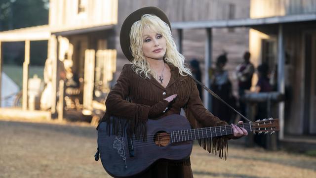 Die Countrysängerin Dolly Parton steht mit Akustikgitarre vor einer Wildwest-Kulisse.