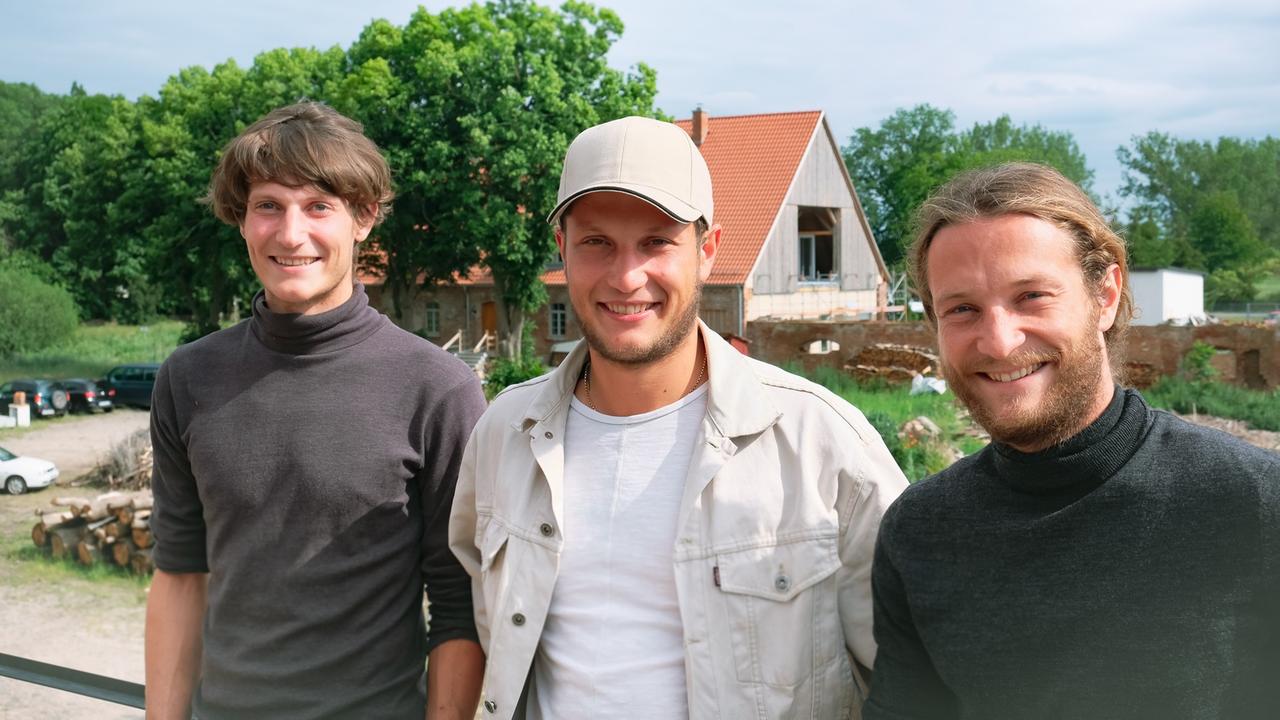 Drei junge Männer stehen nebeinander, im Hintergrund ist ein Haus im Bau zu sehen.