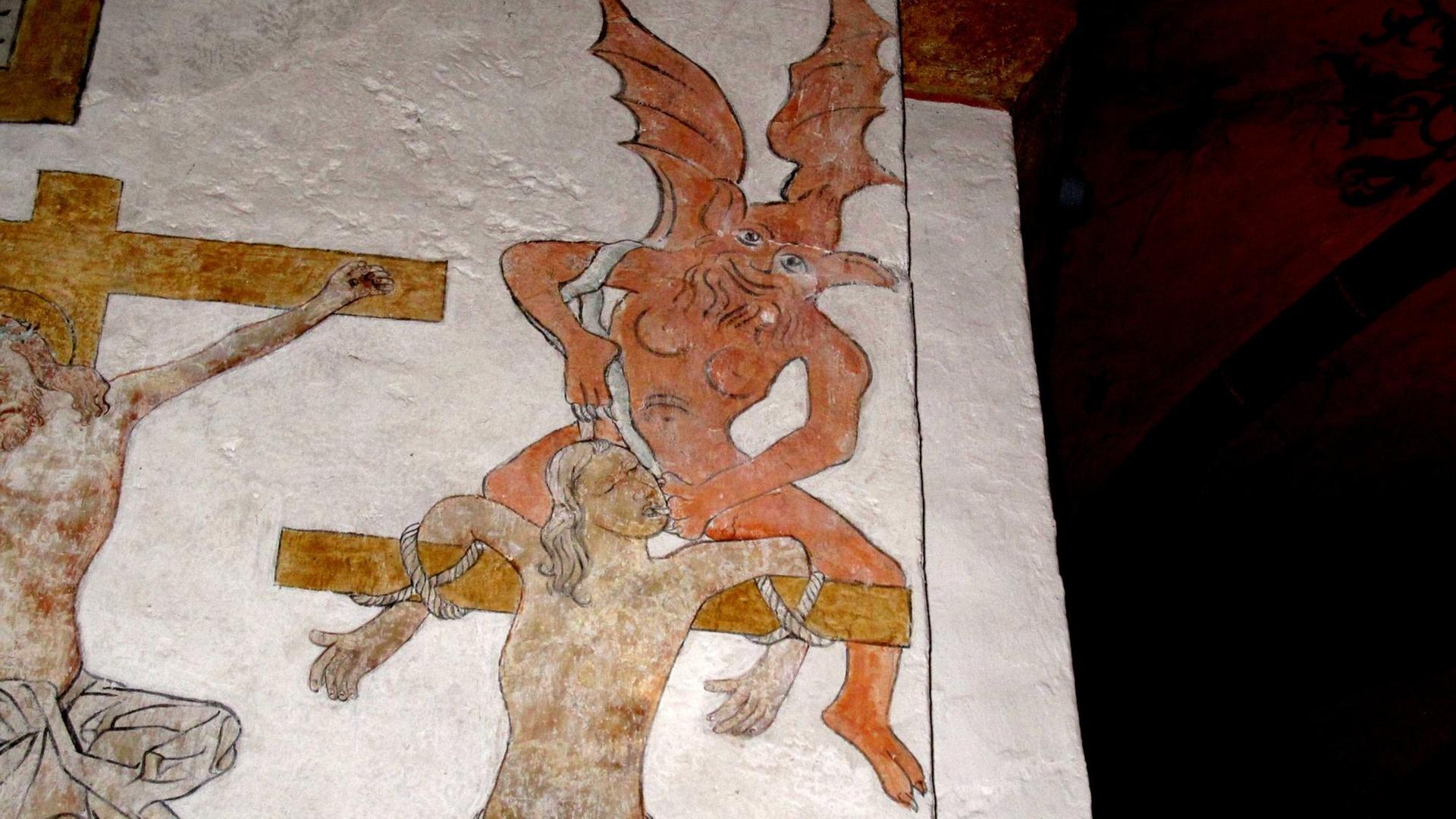 Ein roter Teufel mit Vollbart, Brüsten und Drachenflügeln beugt sich über einen Gekreuzigten. Ein roter Teufel mit Vollbart, Brüsten und Drachenflügeln beugt sich über einen Gekreuzigten.