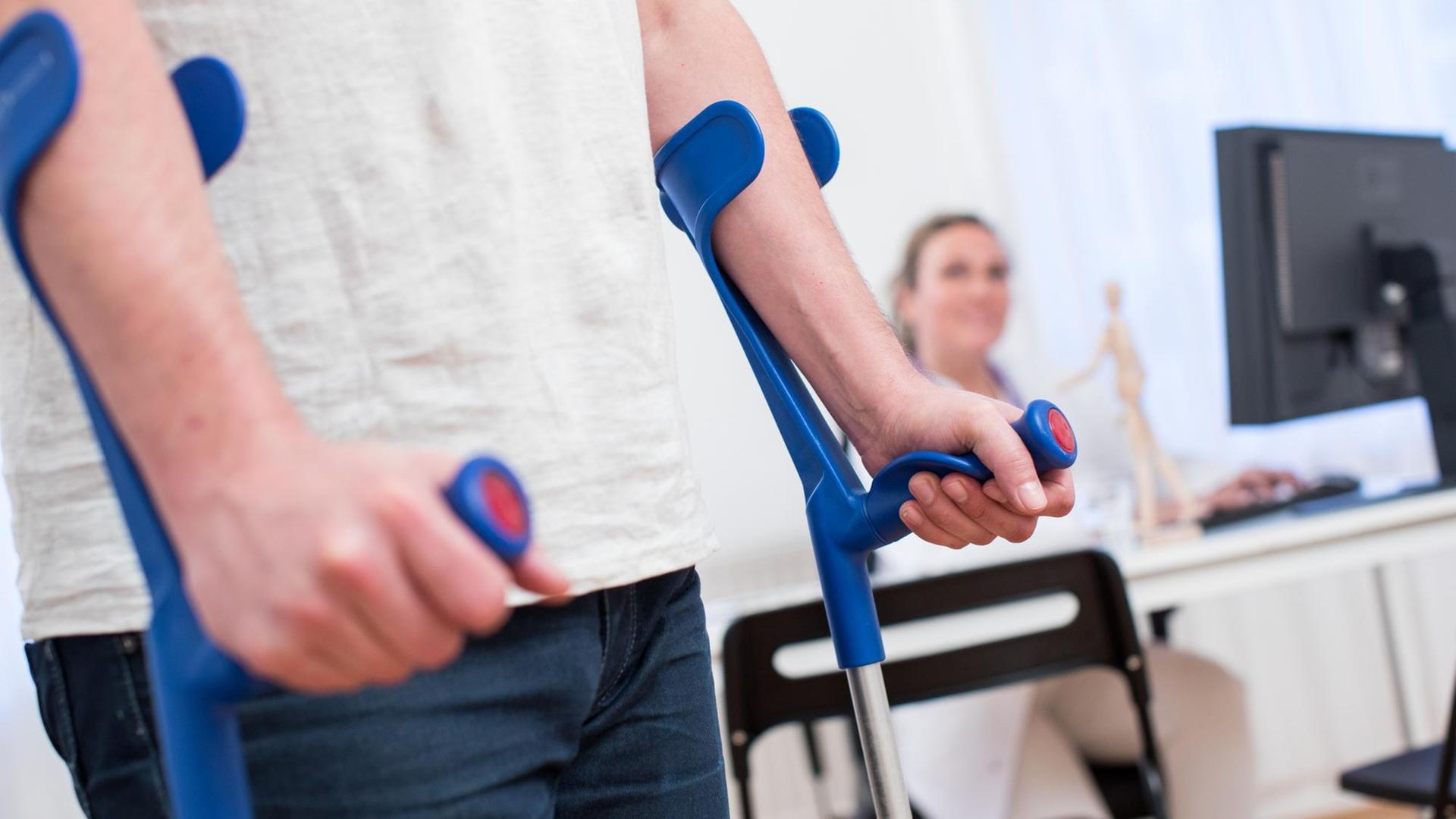 Eine Ärztin sitzt am in einer Arztpraxis am Schreibtisch, während ein Patient mit Krücken im Sprechzimmer steht. Eine Ärztin sitzt am in einer Arztpraxis am Schreibtisch, während ein Patient mit Krücken im Sprechzimmer steht.