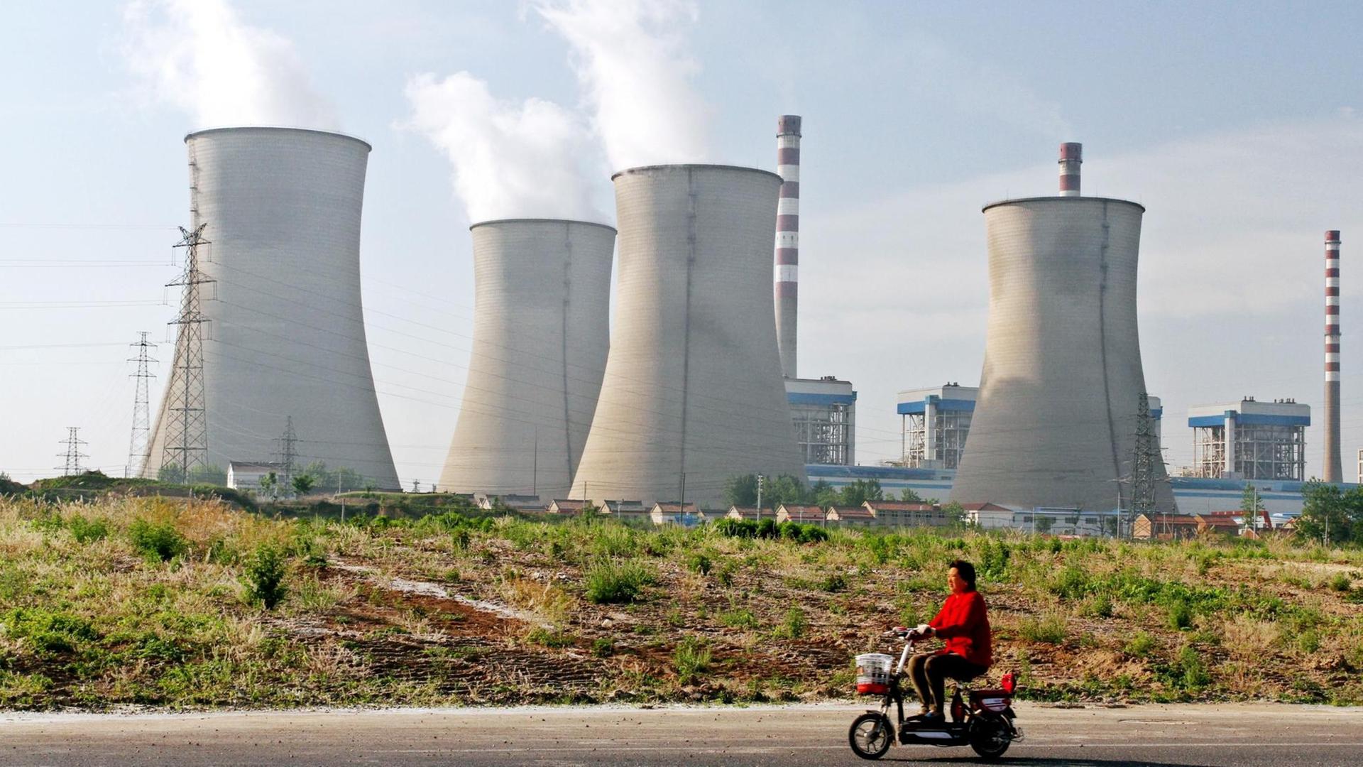 Eine Frau auf einem Elekrofahrrad fährt an einem Kohlekraftwerk in China, Huaian city, Jiangsu vorbei