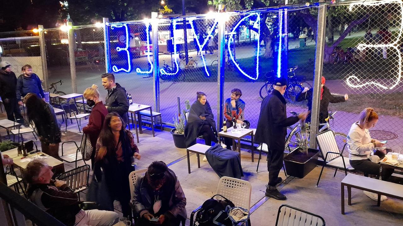 Im Hof des Clubs sitzen viele Mensche in Jacken und Decken gehüllt an Tischen und nehmen Getränke zu sich. Am Zaun steht in Neonbuchstaben "Idylle" geschrieben. Im Hof des Clubs sitzen viele Mensche in Jacken und Decken gehüllt an Tischen und nehmen Getränke zu sich. Am Zaun steht in Neonbuchstaben "Idylle" geschrieben.