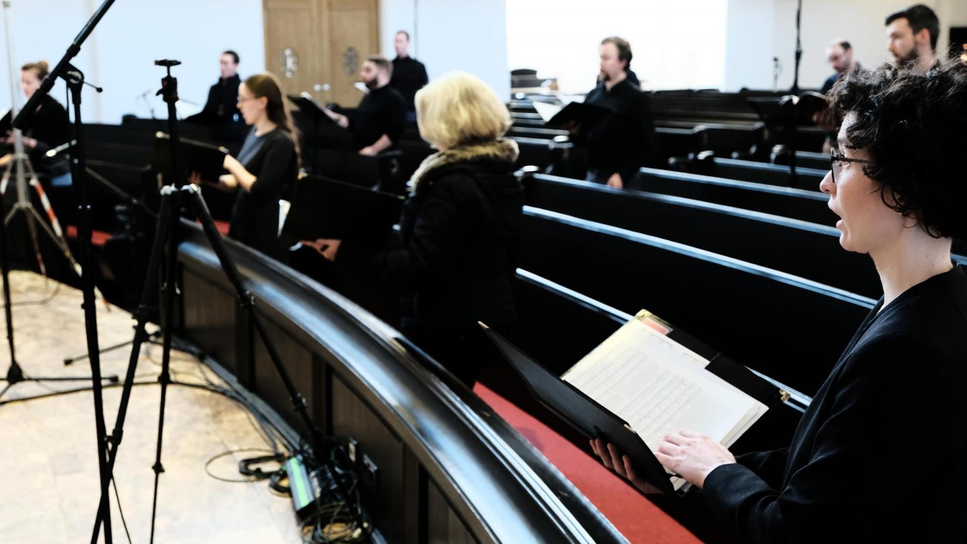 Im Vordergrund ist eine schwarz gekleidete Sängerin zu sehen, die in einer dunklen Kirchenbank steht. Im Vordergrund ist eine schwarz gekleidete Sängerin zu sehen, die in einer dunklen Kirchenbank steht.