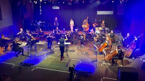 Auf einer Bühne spielt ein Kammerorchester, davor gibt eine Dirigentin einen Einsatz. Auf einem Podest im Hintergrund stehen weitere Musiker, darunter ein Schlagzeuger und zwei Sängerinnen. Scheinwerfer tauchen die Szene in blaues Licht.