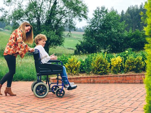 Eine jüngere Frau schiebt auf einer Gartenterrasse einen Rollstuhl, in dem eine ältere Frau sitzt.