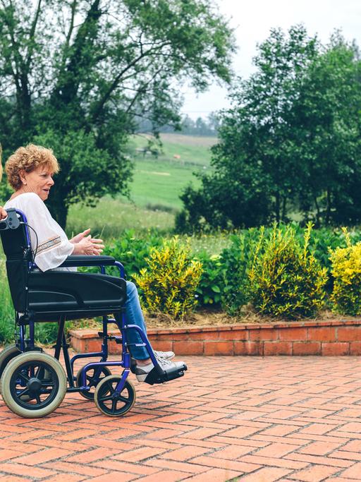 Eine jüngere Frau schiebt auf einer Gartenterrasse einen Rollstuhl, in dem eine ältere Frau sitzt.