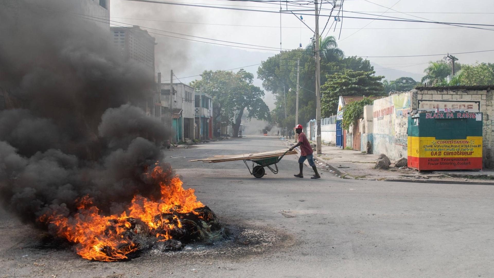 Eine brennende Barrikade in Haitis Hauptstadt Port-au-Prince. Dahinter geht eine Person mit einer Schubkarre die Straße entlang. Eine brennende Barrikade in Haitis Hauptstadt Port-au-Prince. Dahinter geht eine Person mit einer Schubkarre die Straße entlang.